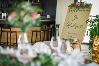 Elegante Hochzeitstafel mit Willkommensschild, floraler Dekoration, stilvollen Gedecken und Barbereich im Hintergrund.