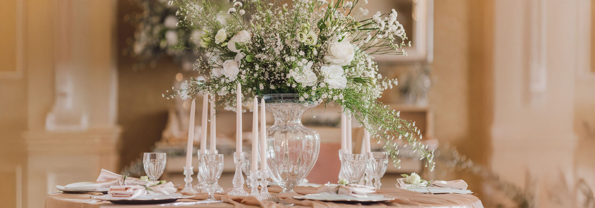 Elegant gedeckter Tisch mit Glasvase voller weißer Blumen, Kerzen und Kristallgläsern für eine Hochzeit