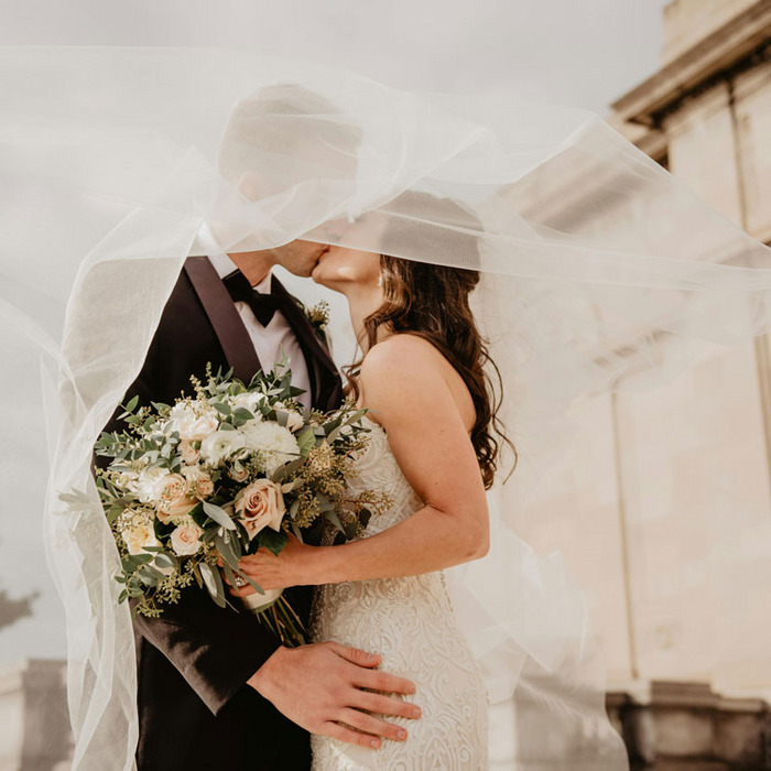 Brautpaar unter Schleier mit Blumenstrauß vor historischer Architektur